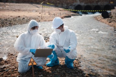 Ekolojist ölü balığı incelemek ve dizüstü bilgisayara veri kaydetmek için örnek alıyor, biyolog koruyucu giysi giyiyor ve endüstriden, sorun ortamından gelen atık su örneklerini maskeliyor