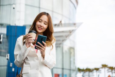 Young business woman smiling holding mobile phone with coffee take away going to work early in morning, Asian businesswoman with smartphone and cup coffee standing against street building near office