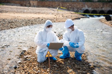 Biyolog koruyucu kıyafet giyer ve maske endüstriden, sorunlu çevreden atık su numunesi toplar, ekolojist örneği incelemek ve veri kaydetmek için ölü balığı laptopa, toksik suya götürür.