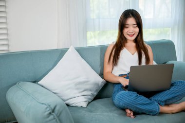 Happy woman typing email on notebook computer, Asian young female smiling sitting relaxing on sofa using laptop in living room at home, freelance browsing through the internet during free time