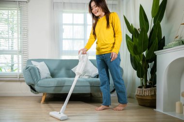 Ev hanımı kadın toz temizleme koltuğun altında ya da koltuk mobilyalarının altında elektrikli süpürgeyle mutlu Asyalı genç kadın evde elektrikli süpürge ile oturma odasında, ev ve ev işi konseptinde.