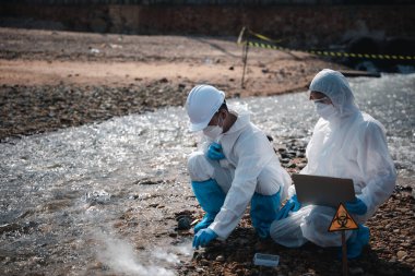 Nehirden çıkan zehirli kimyasalları test tüpü camlarıyla test eden ve beyaz duman çıkaran ekolojist, koruyucu kıyafet giyiyor ve maske endüstriyel, sorunlu ortamdan örnek su topluyor.