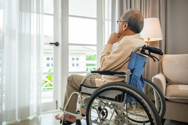 Asian senior man disabled sitting alone in wheelchair looking through window at hospital, lonely elder thoughtful sad old man look outside windows in bedroom at retirement home, feel depressed lonely