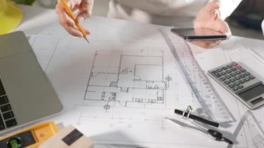 Mimari proje işyeri. Mimar, mimarlık bürosundaki masa masasındaki plan taslağında kalemle çizim yapmadan önce ev tasarımını gözden geçirmek için akıllı telefon veya cep telefonu kullanır