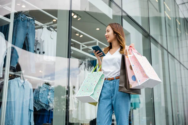 Asian beautiful woman holding shopping bag and smartphone on her hands near the mall shop window, Smiling shopper young girl standing using mobile phone after shopping on city street