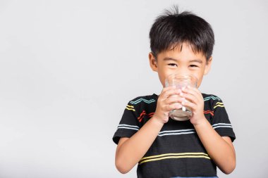 5-6 yaşlarında küçük tatlı bir çocuk beyaz arka planda izole edilmiş beyaz çekimde camdan tatlı su içerek gülümsüyor. Asyalı çocuklar anaokulu, günlük yaşam sağlığı.