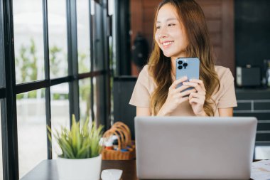 Happy lifestyle female smiling using smartphone in coffee shop, young business woman working with laptop computer she holding smart mobile phone looking out of windows at cefa for texting messages