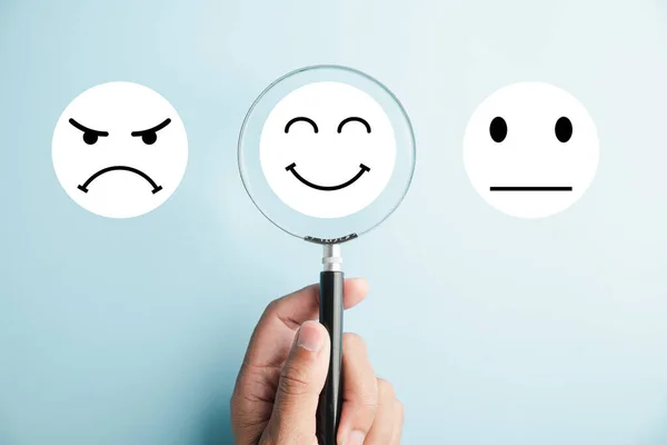 stock image Hand holds magnifier, emphasizing happy smiley face icon among emotions. Good feedback rating and positive customer review. Experience, satisfaction survey, mental health. signifies satisfaction.