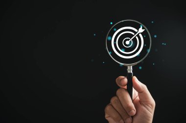 Büyüteçle incelenen bir hedef tahtası özellikleri, iş amaçlarına odaklanılmasını vurguluyor. Bir arka plan karşısında, arama, hedefe ulaşma ve stratejik başarı kavramlarını temsil eder..