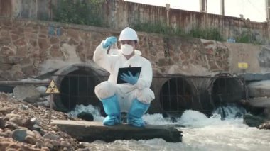 Nehir kontrol listesinden zehirli kimyasallardan oluşan suyu örnekleyen ve pano kağıdına yazan ekolojist, koruyucu kıyafet giyiyor ve endüstriyel su örneklerini maskeliyor.