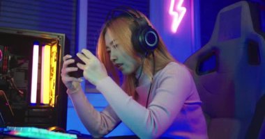 You win. Excited Asian woman gamer wearing gaming headphones playing joystick console video game and streaming online on computer PC neon light at home happy successful, tournament player E-Sport