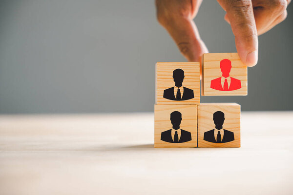 A hand is seen choosing man people icons with on cube wooden toy blocks stacked. Represents the idea of teamwork, leadership, and corporate strategy in achieving business goals.