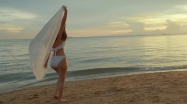 Güzel, genç bir kadın rüzgarda bikini mayo giyer, eşarp takar, tropikal plajlarda yürür ve altın gün batımında özgürlük için elini kaldırır.