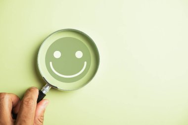 Elle tutulan büyüteç, gülen surat ikonunu seçiyor. İyi geri bildirim notu, olumlu müşteri değerlendirmesi. Tecrübe, tatmin zihinsel sağlık araştırması. Sertifika memnuniyeti temsil eder