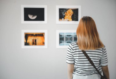 Asian young woman standing she looking art gallery in front of colorful framed paintings pictures on white wall, female watch at photo frame to leaning against at exhibit museum, Back view