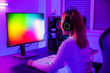 Asian professional gamer playing online video game on desktop computer PC have colorful neon LED lights dark room, young woman in gaming headphones using computer for playing game at home, Back view