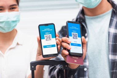 Koronavirüs salgını sırasında akıllı telefon ekranında dijital aşı sağlık pasaport uygulamasını gösteren Asyalı genç kadın ve erkek seyahat etmeye hazır.