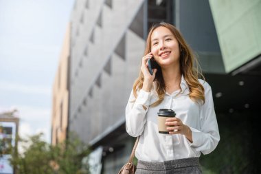 Asyalı genç iş kadını dışarıda cep telefonuyla konuşuyor kahve fincanı servisi yapıyor, yaşam tarzı iş kadını elinde kağıt bardak tutuyor ve ofisin dışındaki akıllı telefonu arıyor.