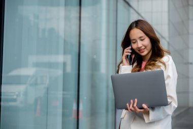 Asian businesswoman working on laptop and talking on cell phone at front building near office, Portrait beautiful business woman in white suit smile discussing issues on smart mobile phone in city