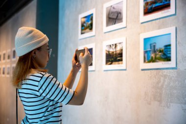 Asyalı kadın önünde resim galerisi koleksiyonu yapıyor. Çerçevelenmiş resimler beyaz duvarda cep telefonlu resimler, insanlar resim çerçevesinde akıllı telefon ile sanat galerisindeki resimleri izliyor.