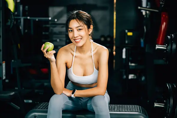 Asian woman holding a green apple in a fitness gym, emphasizing the importance of clean, healthy eating as part of a workout routine. Healthy fitness and eating lifestyle concept