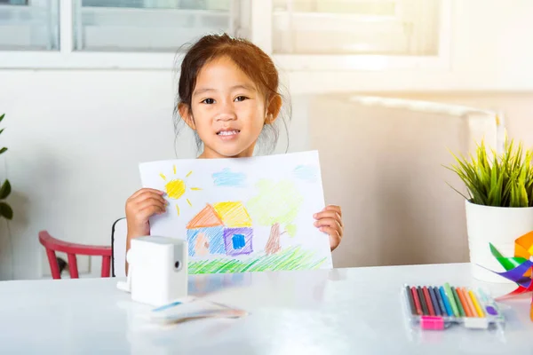 Happy child little girl lift up colorful drawing landscape my home dream on paper to camera, Asian cute kid preschooler smiling showing draw country house picture at home to learning arts homework
