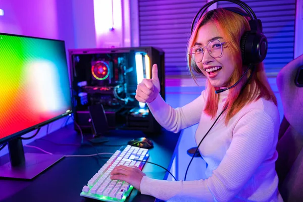 Gamer playing online game wear gaming headphones looking to camera expressing success with game giving thumbs up sign, Smiling woman live stream she play video game at home neon lights living room
