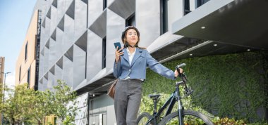 Happy lifestyle business female commuting outside in morning, Asian smiling young woman use smart mobile phone talk with business during go to office work at city street with bicycle, Eco friendly