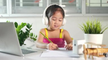 Asyalı çocuk evde video konferansı eğitimi alıyor. Küçük çocuk anaokulunda kulaklık takıyor masada oturuyor dizüstü bilgisayar kullanıyor ve öğretmeniyle internet üzerinden iletişim kuruyor.