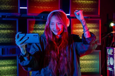 Kazanmak. Oyun odasında heyecanlı hisseden kafası karışmış Asyalı kadın, neon ışıklı akıllı cep telefonuyla video oyunu oynayan Happy Gamer, kutlama yapmak için elini kaldırıyor.