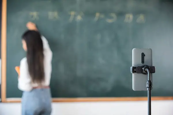 Back to school online class. Asian teacher teach on board while have blackboard lecture online through smartphone in classroom, woman standing front of class live video call online smart mobile phone