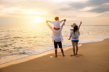 Tatilde mutlu bir aile. Baba, anne ve çocuklar yaz plajında omuz omuza gezintisi, gün batımında çocukları sırtında taşıyan aileler, yaz tatilinde aile.