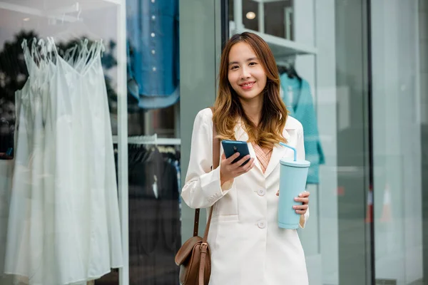 Young Asian woman multitasking in the city ,holding a tumbler mug and smartphone in front of a store window. Digital connectivity and convenience is her lifestyle.