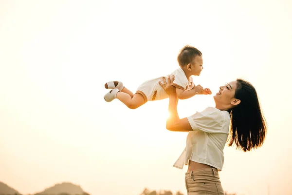 In a playful childhood moment, a happy Asian mom throws her little boy up into the sunny sky. The cheerful child enjoys the freedom of flying, while the joyful mother looks on with happiness and love
