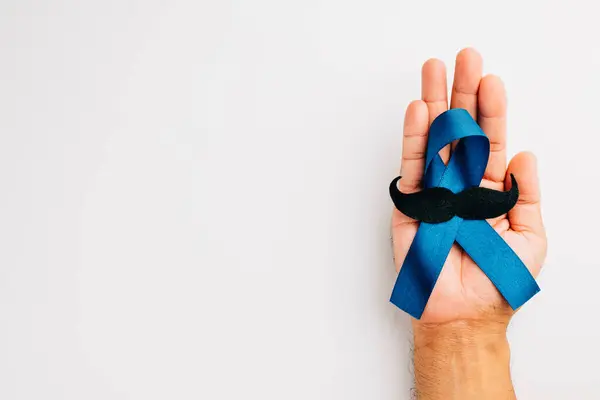 Supporting mens health and Prostate cancer awareness in November. Mans hands hold a light blue ribbon with a mustache on a blue background a symbol of strength and solidarity.