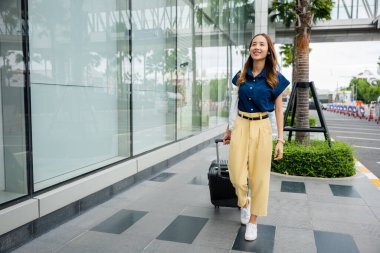 Genç bir turist güneşli bir günün tadını çıkarıyor, valiziyle yürüyor ve akıllı telefonuyla bağlantıda kalıyor..