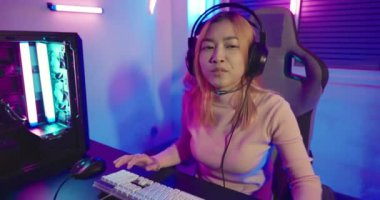 Live stream. Happy gamer recording and saying hello to her online fans during live streaming before playing video game online with computer neon lights, Asian woman wearing gaming headphones at home