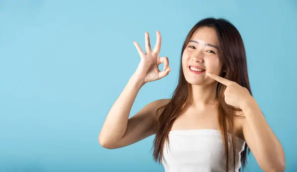 Beautiful female point fingers to smiling mouth and showing OK sign for good studio shot isolated on blue background, Asian young woman pointing to her strong white teeth, Dental health concept