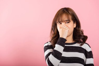 Portrait of an Asian woman, unhappy with a disgusted expression, pinches her nose due to a foul smell. Studio shot isolated on pink, illustrating a healthcare concept. clipart