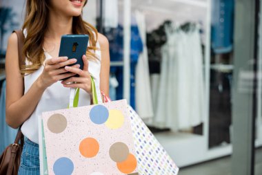 Alışveriş torbaları ve cep telefonuyla birden fazla iş yapan şık bir kadın online alışverişin keyfini çıkarıyor. Giderken son moda ve aksesuarlara göz atıyor..