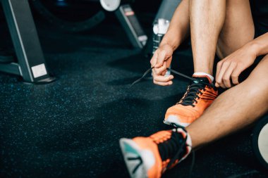 Formda ve kaslı bir erkek spor salonunda bağcıklarını bağlıyor. Etrafı halterler ve diğer spor aletleriyle çevrili. İğne, vücut geliştirme ve vücut geliştirmeye olan bağlılığını vurguluyor..