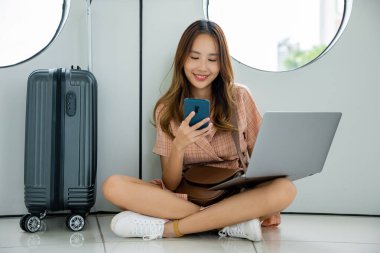 Kadın bavulu ve akıllı telefonuyla rahatça yerde oturuyor ve havaalanında tatilinin tadını çıkarıyor. Turist yolculuğu konsepti