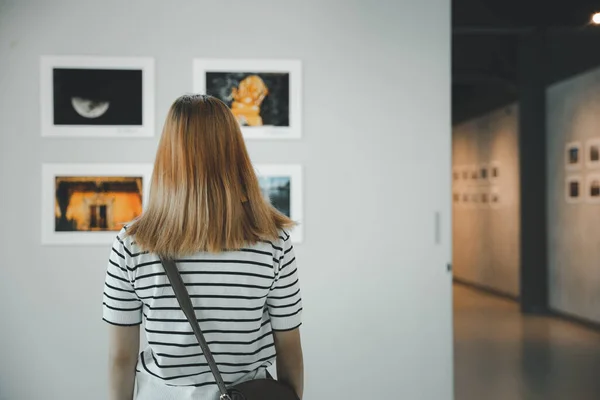 Asian young woman standing she looking art gallery in front of colorful framed paintings pictures on white wall, female watch at photo frame to leaning against at exhibit museum, Back view