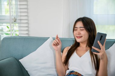 Yaşam tarzı, kablosuz kulaklıklı bir kadın müzik dinlemeli bir şarkı ve müzik uygulamalı akıllı telefondan biraz dans, Asyalı bir kadın oturma odasında akıllı cep telefonuyla kanepede müzik dinlerken kulaklık takıyor.