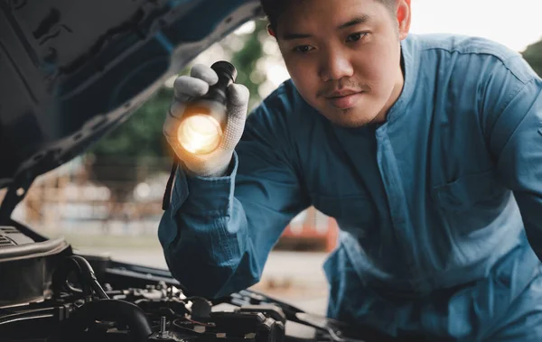 Repairman checking and repairing auto engine with the help of an electric lamp. Asian man mechanic in a blue suit is visible, analyzing car problems on the cradle.