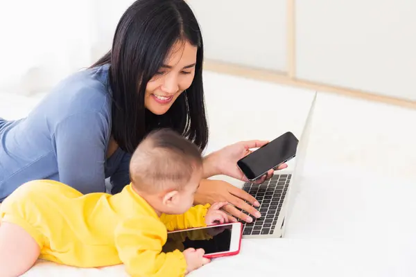 Asian young mother work from home busy on laptop computer and his baby daughter playing smartphone, Freelance career for a young mom