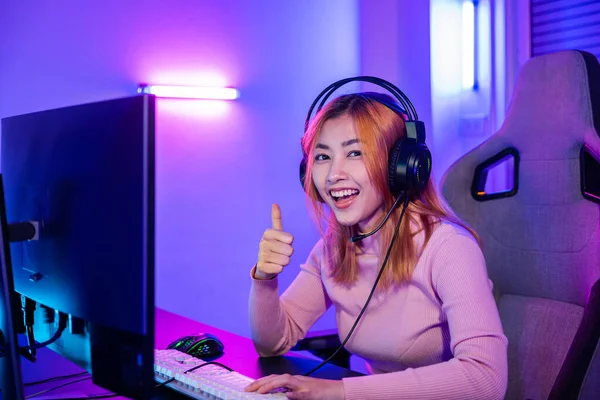 Gamer playing online game wear gaming headphones looking to camera expressing success with game giving thumbs up sign, Smiling woman live stream she play video game at home neon lights living room