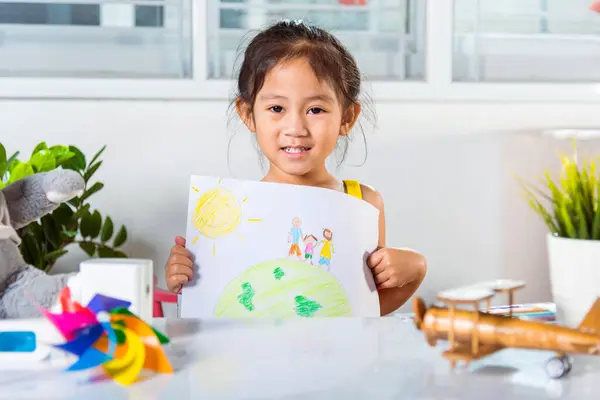 Happy child little girl lift up colorful drawing family standing hold hands on planet earth on paper to camera, Asian cute kid preschool smiling showing draw picture at home earth day concept