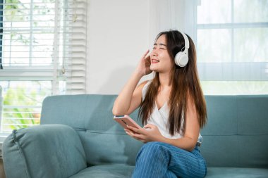 Kulaklık takan Asyalı genç bir kadın akıllı cep telefonuyla müzik dinliyor ve evdeki kanepede otururken dinleniyor. Kadın kulaklık takıyor ve akıllı telefondan müzik dinliyor.