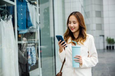 Akıllı cihaz ve sıcak içecek. Elinde bardak bardak ve akıllı telefon tutan genç bir kadın. Dijital bağlantı ve kafein takviyesi onun kolaylığı..
